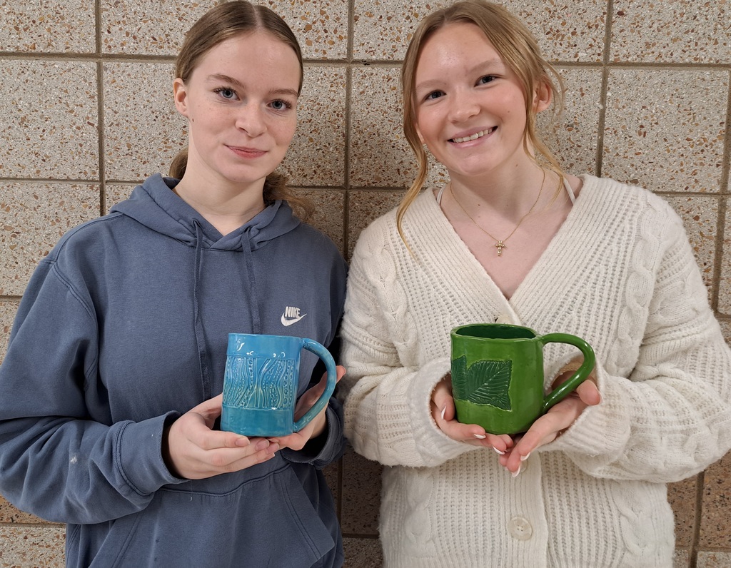 two females, one holding a blue stamped mug and the other holding green stamped mug made out of clay