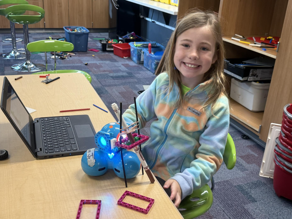 female 2nd grader sitting at her desk with her robot that is holding markers that she coded to draw