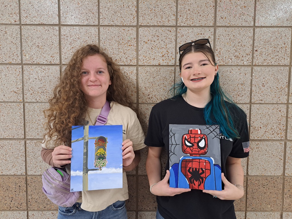 two female students holding paintings, one of spider man