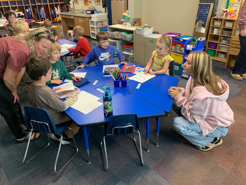 Kindergarten students sitting at their table with a teacher and a HS student helping them