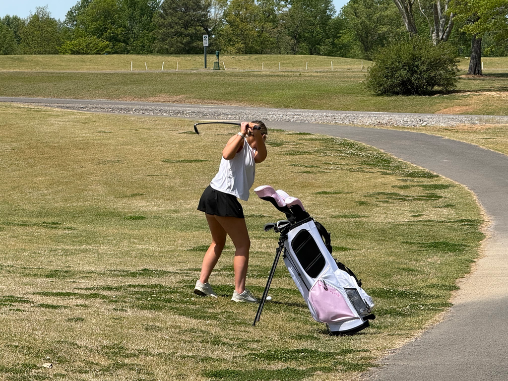 Person with a golf club, standing on grass near a pathway with a golf cart. Background shows trees and a grassy area.