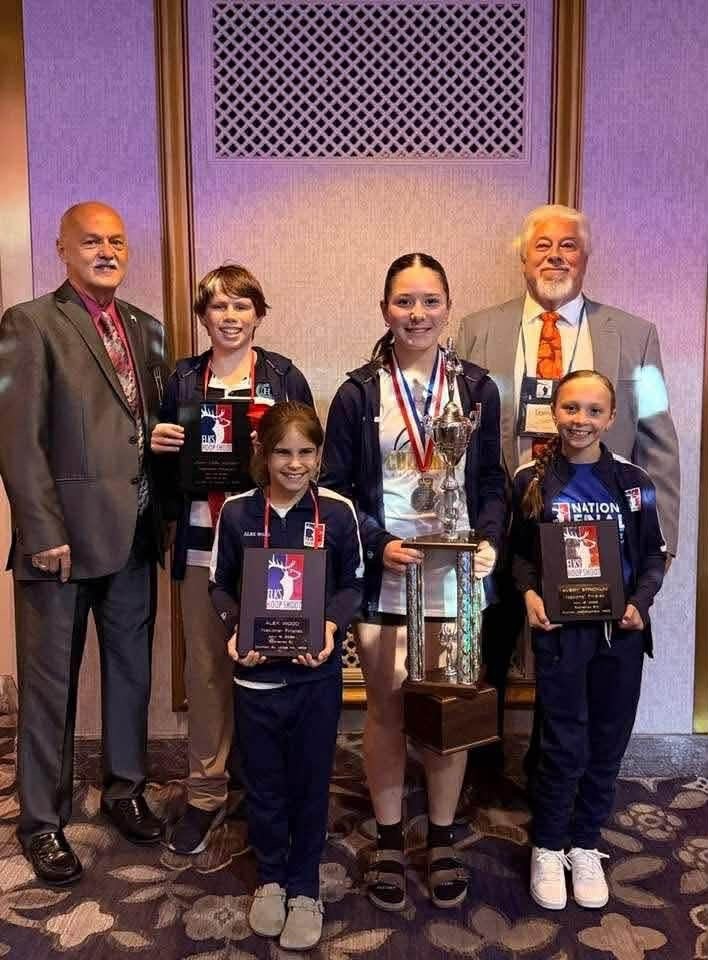 Six children, one adult, and two men in suits. All pose with medals and trophies. Purple wall in background.
