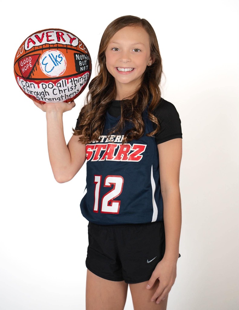 A girl in a basketball jersey poses with a ball, smiling and holding it up.