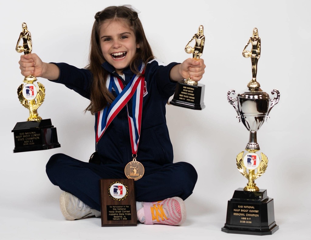 A girl in a navy blue outfit is seated, holding three trophies and a medal. She is smiling.