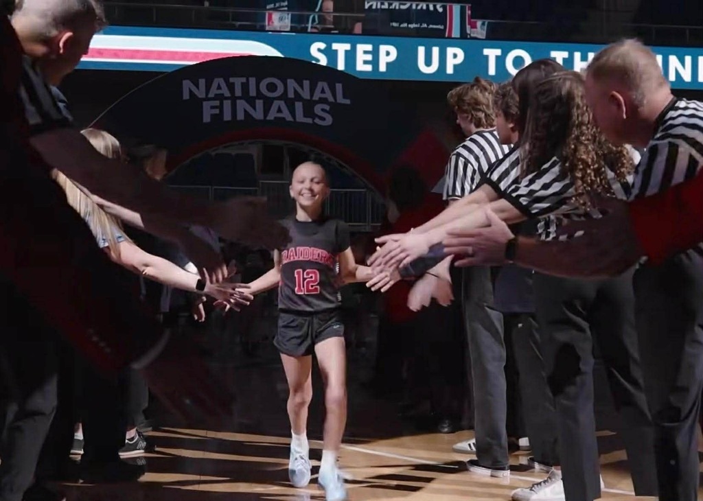 A young girl in a black jersey walks across a basketball court as people in striped shirts and black pants surround her.