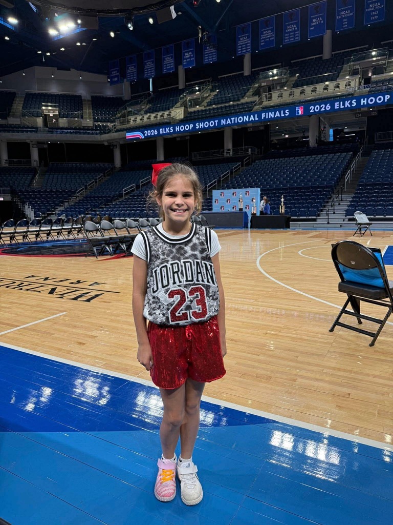 A young girl stands on a basketball court, wearing a red and black jersey. A blue floor is visible, and an empty bleachers is in the background.