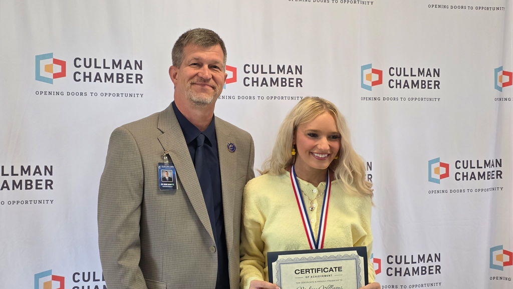 A man and woman stand together at an awards ceremony. The woman holds a certificate and both wear formal attire.