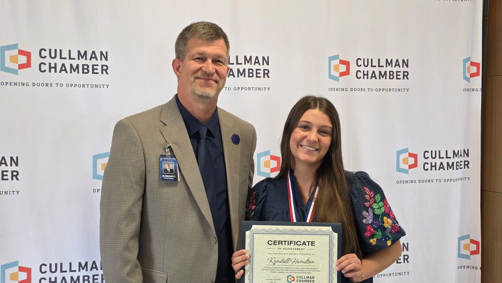 A man and woman stand smiling, holding a certificate. The man wears a suit and the woman a floral shirt. Behind them, a Cullman Chamber banner displays.