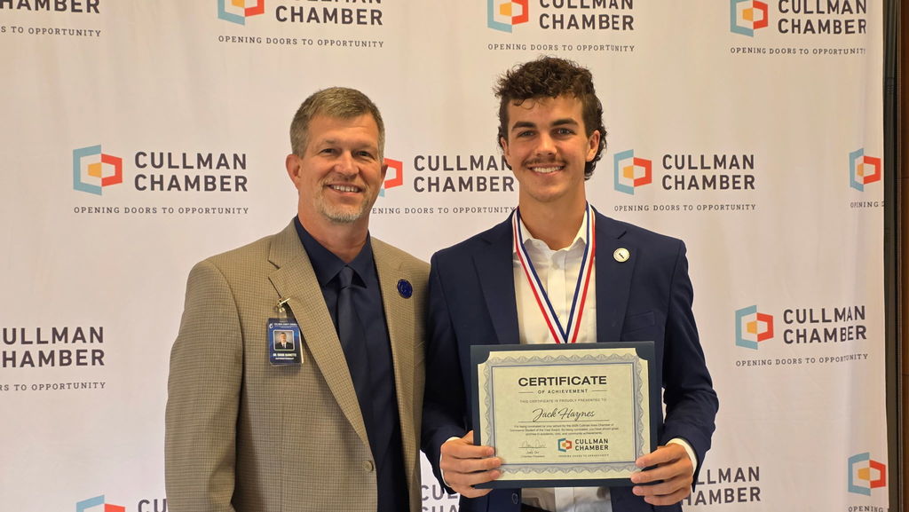 Two men stand next to each other in front of a banner, one holding a certificate.