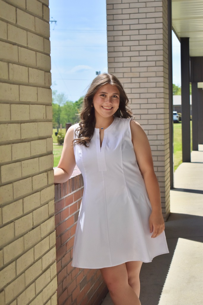 A woman in a white dress stands in a corridor, leaning against a brick wall with a grassy area visible.
