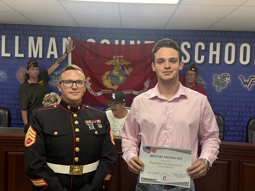 Two men stand next to each other holding a certificate. One wears a Marine uniform, and the other a pink shirt. A flag and text reading "SCHOOL" are behind them.