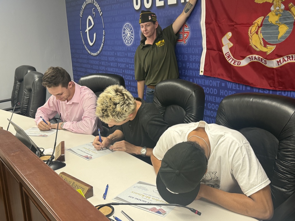 Three people are seated at a table signing documents; one is wearing a cap, one has a watch, and a Marine flag is in the background.