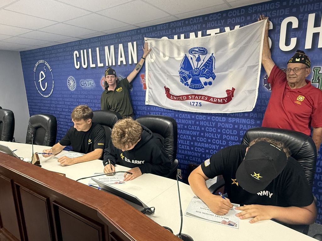 People seated around a desk hold a flag and a banner reading "Cullman County". They are signing papers.