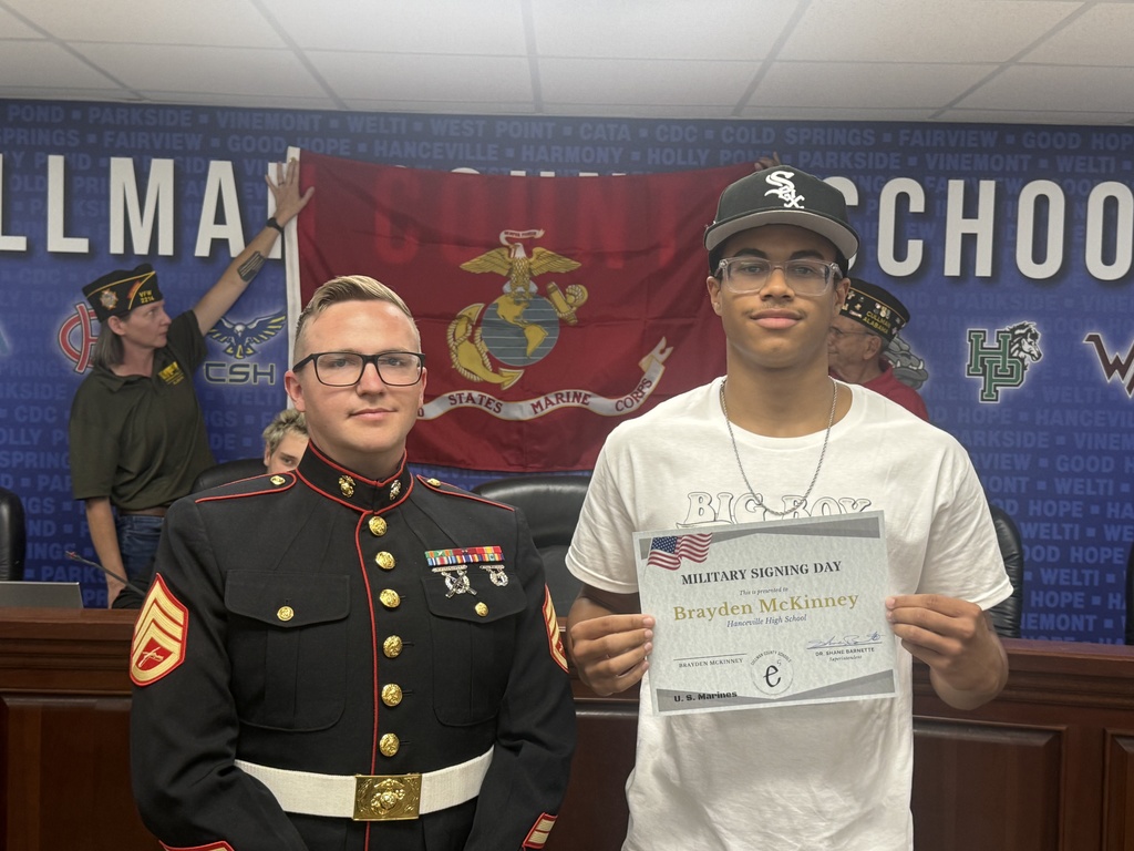 Two men in uniform, one in a military uniform and the other in a white shirt, stand in front of a wall with a flag and a sign.