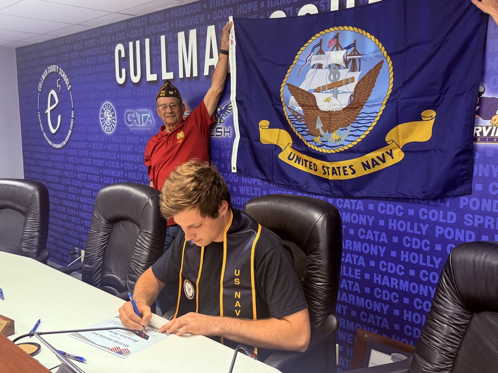 Two men in a room with navy-themed decor. One man signs a document at a table, while another stands and holds a flag.