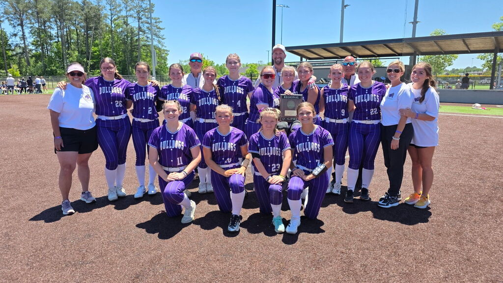 A softball team poses on a field with two adults, one holding a trophy, under a clear sky.