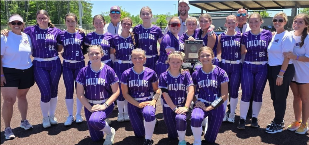 A softball team in purple uniforms poses with an award on a field with a shelter.