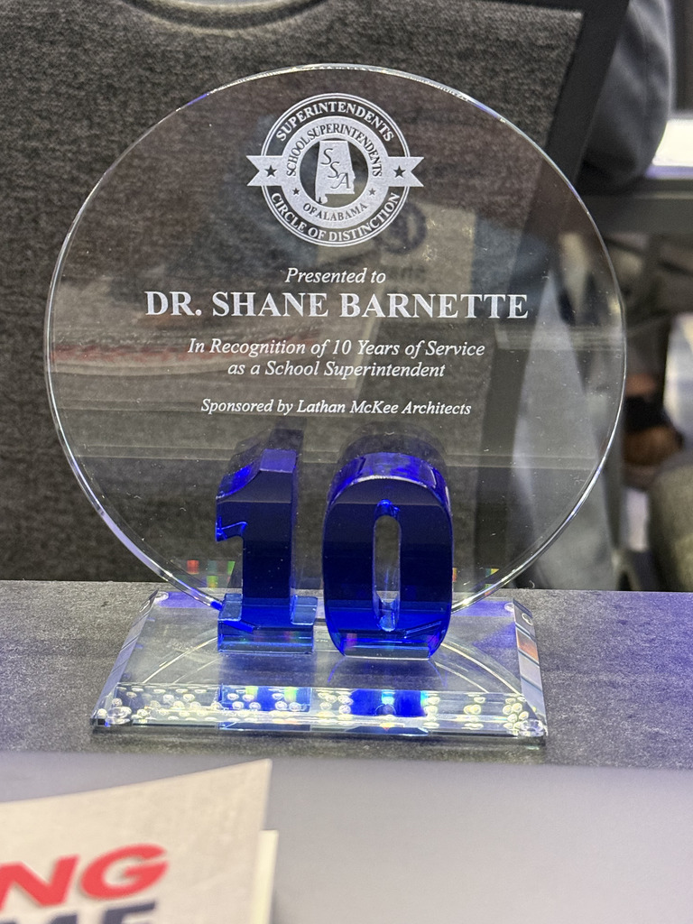 A round blue plaque with "10" in the center, honoring Dr. Shane Barnette for 10 years of service as a school superintendent.