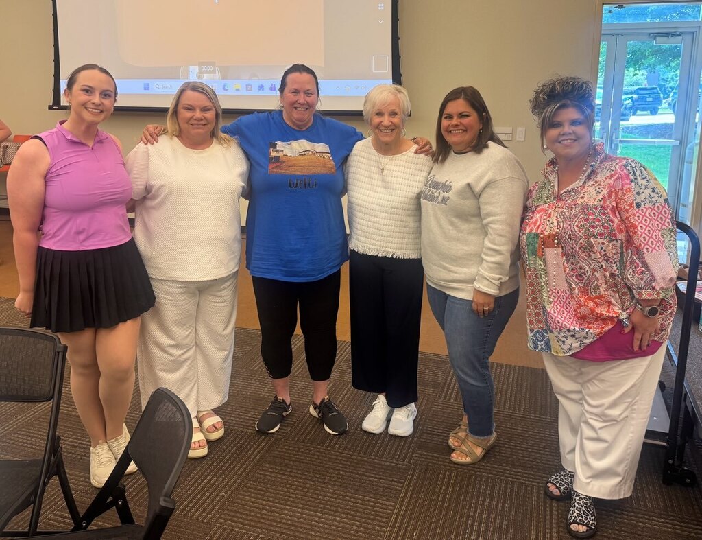 Six women in casual clothing standing close together, smiling. Behind them, a projector screen is visible.