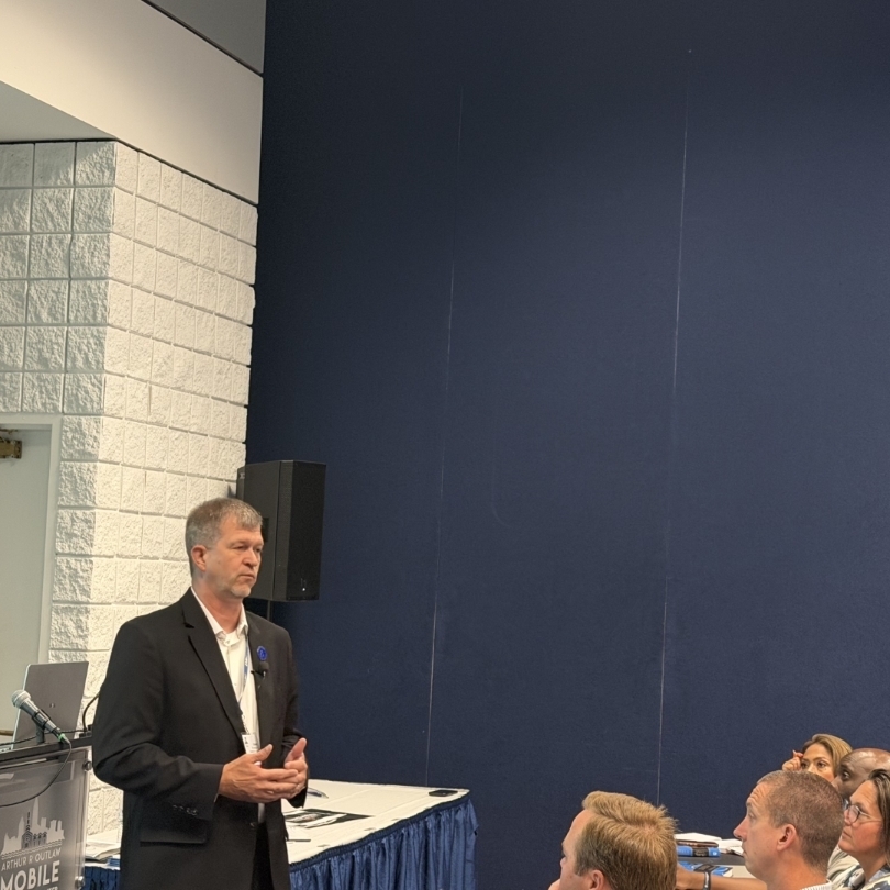 A man in a suit addresses a seated audience. Behind him, a blue wall contrasts with a white-tiled section.
