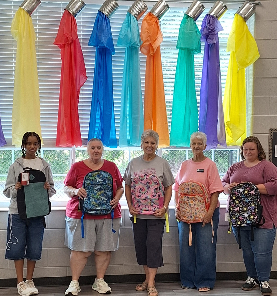 Five adults stand in front of a window, each holding a backpack. Colorful plastic bags hang above.