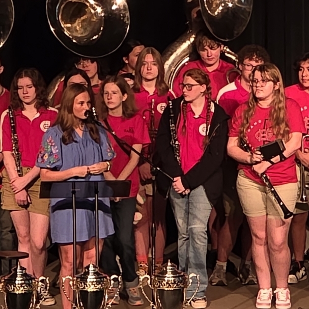 A group of musicians with instruments on stage, some standing, others holding instruments, a woman in front speaking.