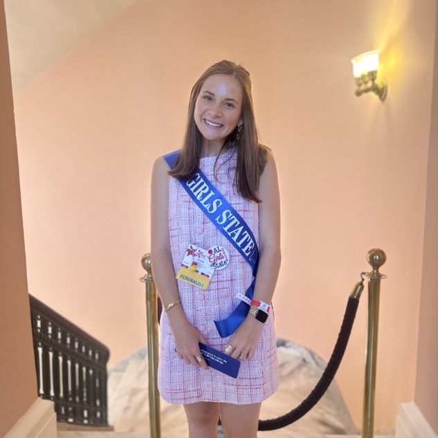 A woman in a pink dress and blue sash stands on a staircase, smiling for the camera.