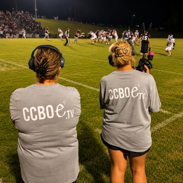 Two women in gray shirts labeled "CCBOE.TV" are watching a football game on a field.