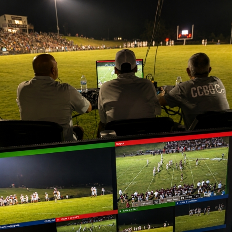 Three people sit in chairs in a stadium, focused on monitors displaying the ongoing football game.