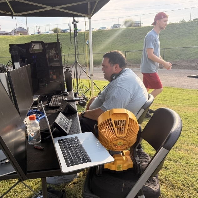 A man sits at a table with multiple computer monitors, a laptop, and various items. Another man stands in a grassy area nearby.