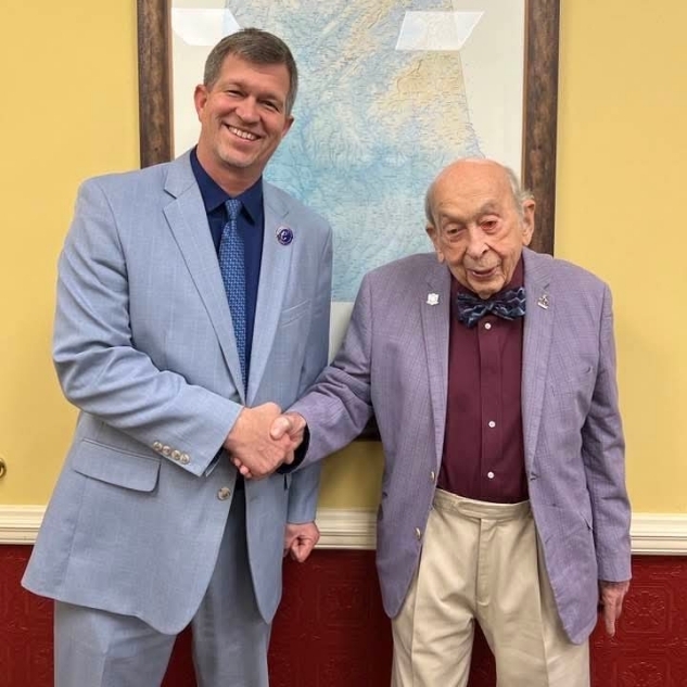 Two men in suits and bow ties stand next to each other, shaking hands. One wears light blue, the other dark purple. A framed map hangs on a yellow wall behind them.