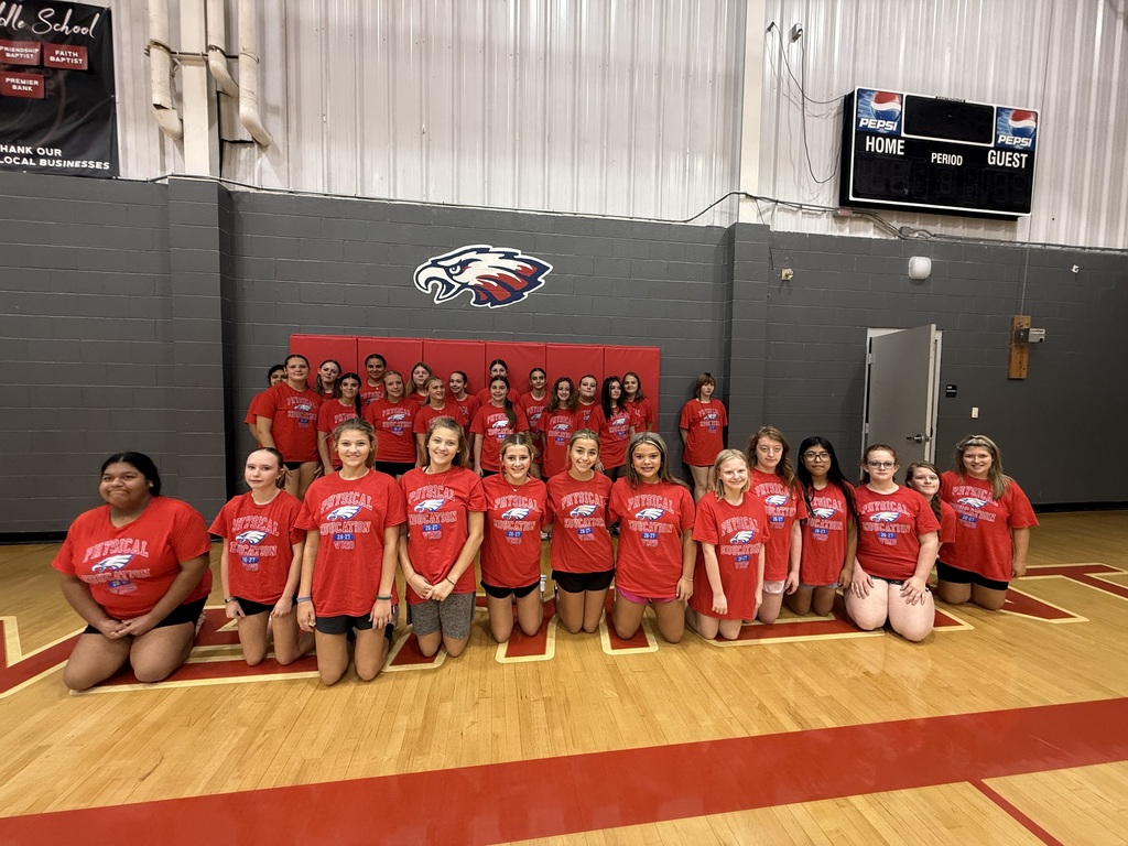 A team in red shirts poses for a photo on a basketball court. Behind them is a red wall with a logo.