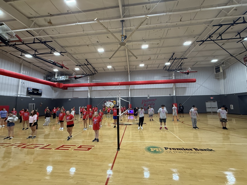 A group of people playing volleyball in an indoor court. They wear various colored jerseys. The floor has a red line and "Eagles" printed.