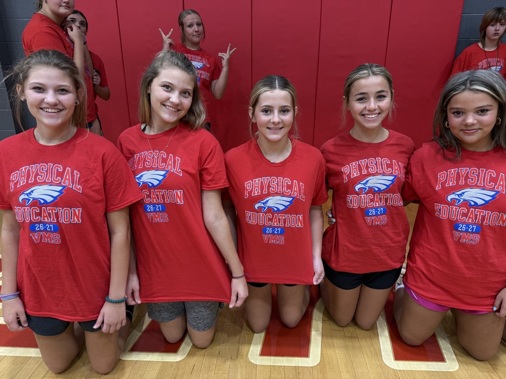 Five girls in red shirts with the word "Patriots" on them stand together in a gym with a red wall.
