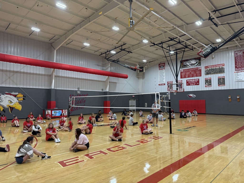 An indoor basketball court has a volleyball net. Several individuals sit on the floor in a circle.