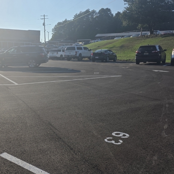 Cars parked in a lot with numbered parking spaces, a building visible in the background, and trees.