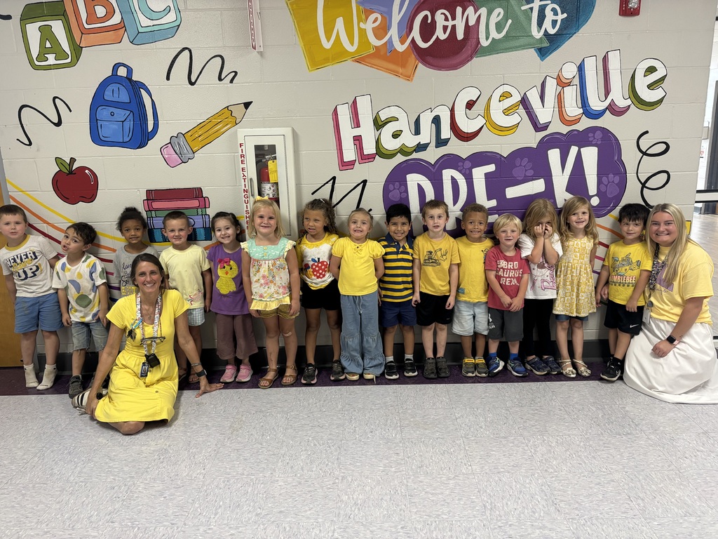 A group of children in yellow shirts and shorts stand before a wall with a colorful mural.