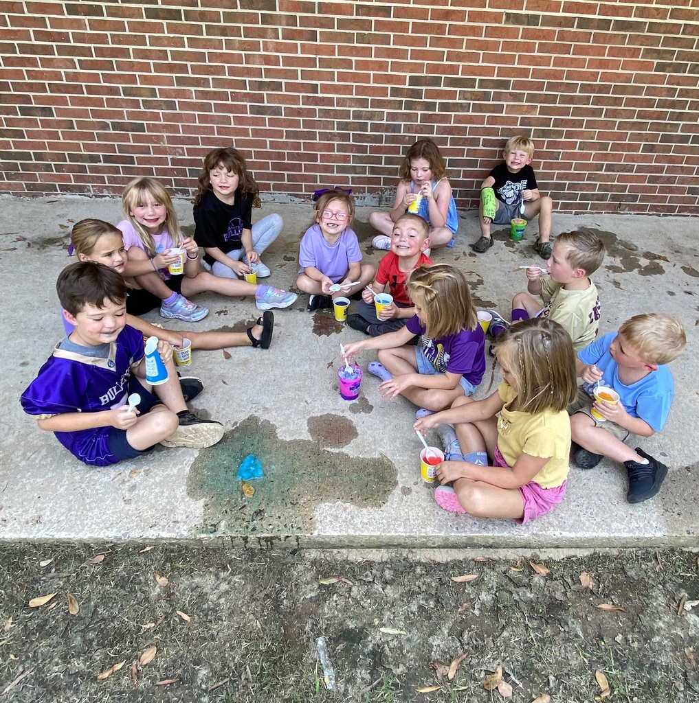 A group of children seated in a circle outside, some holding cups, in front of a brick wall.