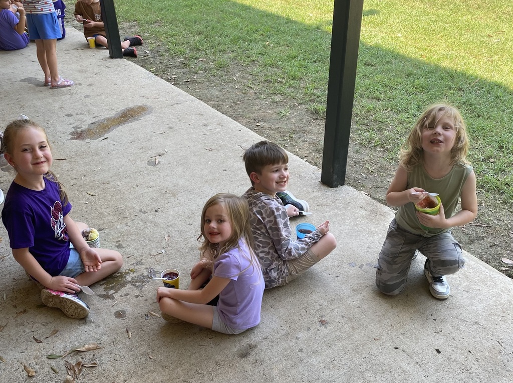 Four children sit on a concrete patio, with one holding a cup, while a grassy area and people are behind them.