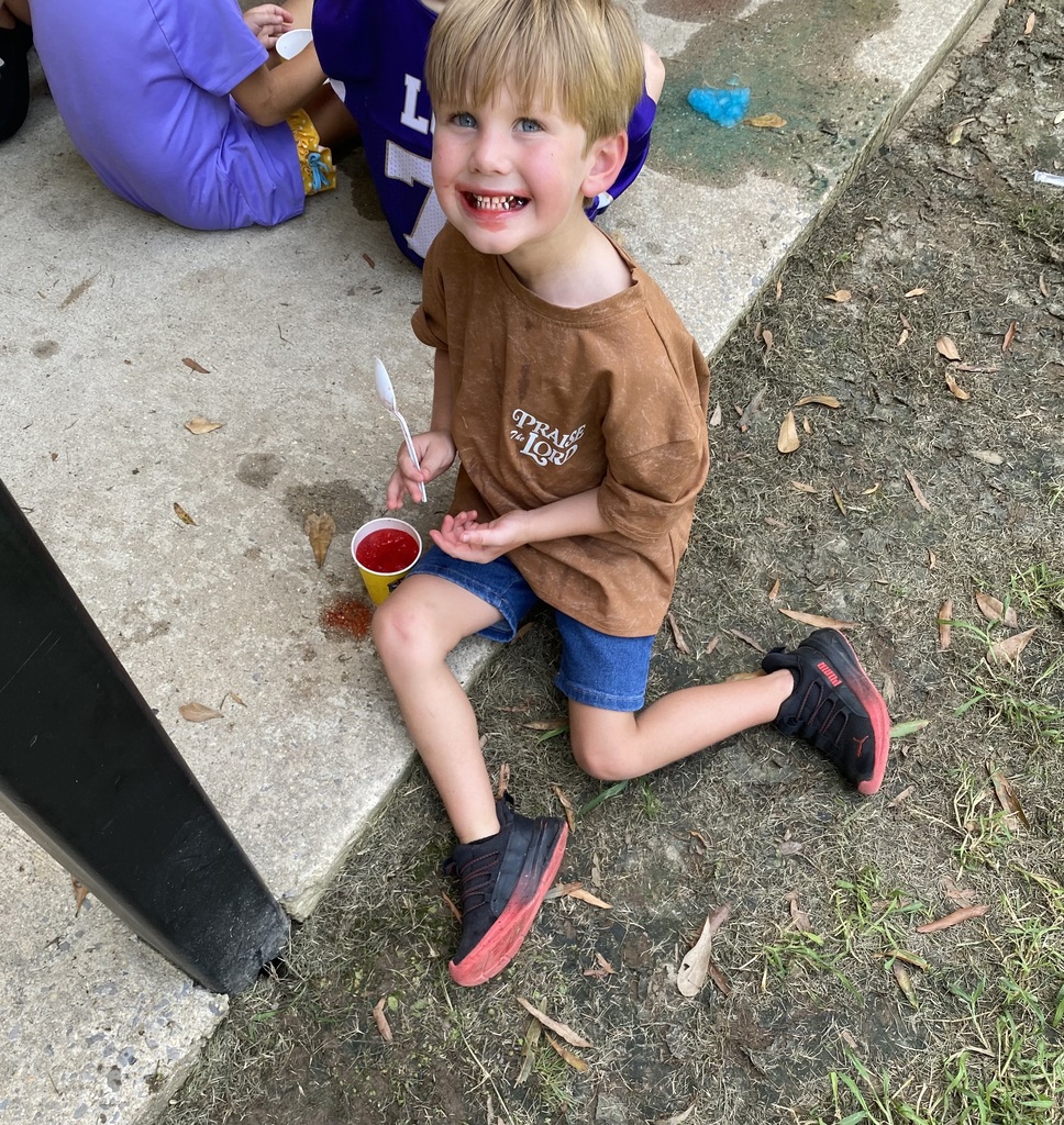 A young boy wearing a brown shirt and denim shorts, sitting on concrete, holding a cup. Another child in a blue shirt sits behind him.