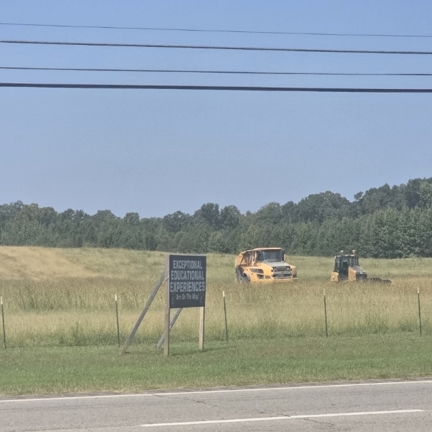 A yellow construction vehicle in a grassy field, with a sign and people nearby, surrounded by trees.