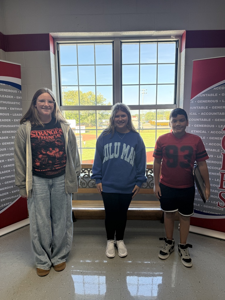 Three people in a room, one wearing a gray sweatshirt, one in blue, and one in red. A window and bench behind them.