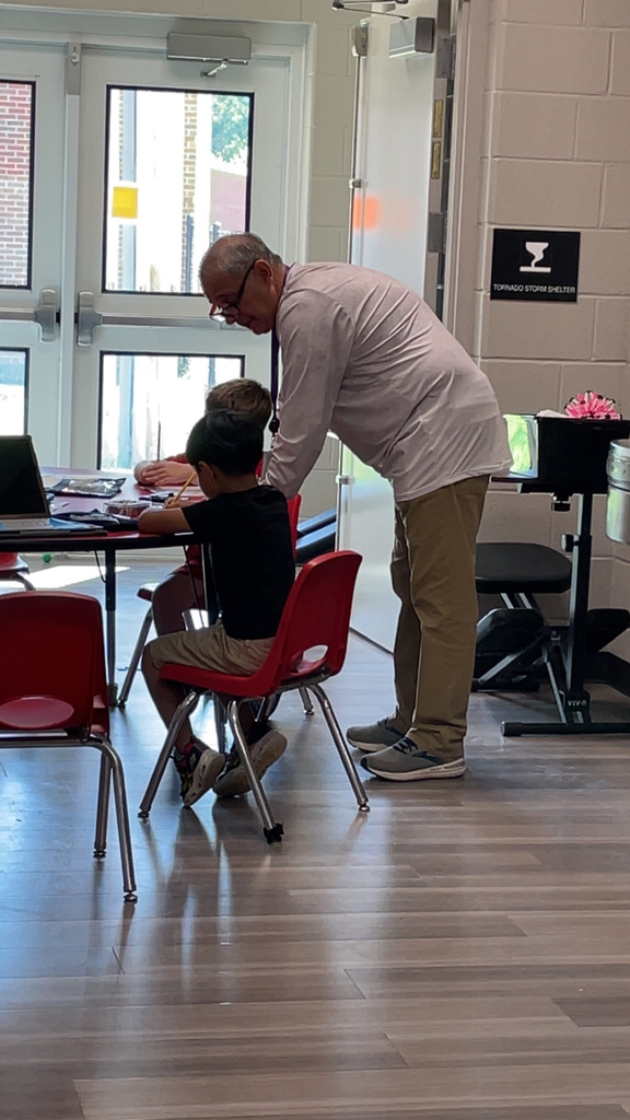 An older man in glasses bends over a table where two children sit. A door with glass panels and a sign are behind them.