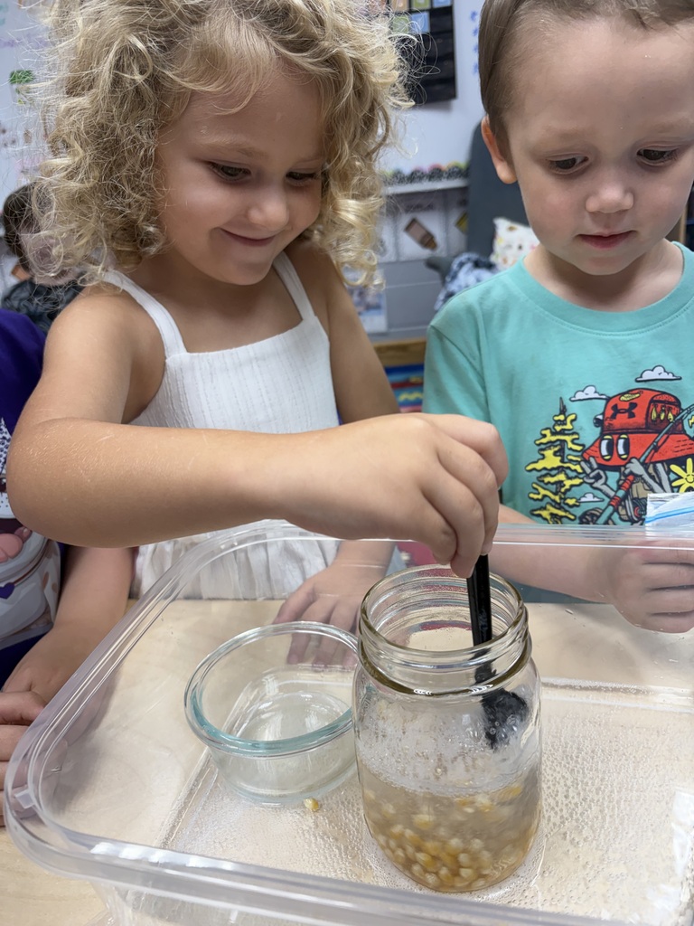 Two children are engaged in a science activity, mixing ingredients in a jar with a stick.