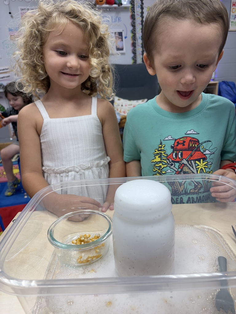 Two children look at a science experiment with a clear plastic container, a bowl, and a bottle.