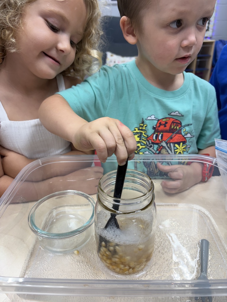 Two children explore a jar with a spoon and beans on a clear tray. One child looks closely at the jar.