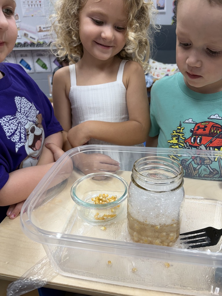 Three children examine a jar with a spoon and fork on a table, with a plastic tray and bowl.