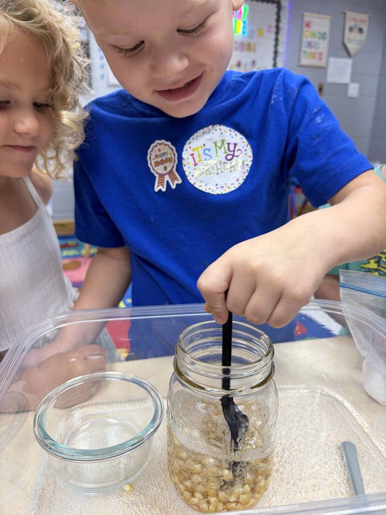 A boy in a blue shirt stirs beans in a clear jar while a girl in a white dress looks on.