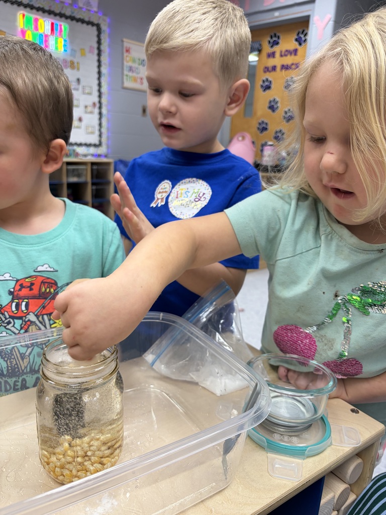 Three children in a classroom pouring corn kernels into a jar. One child has light blonde hair, another has dark brown hair.