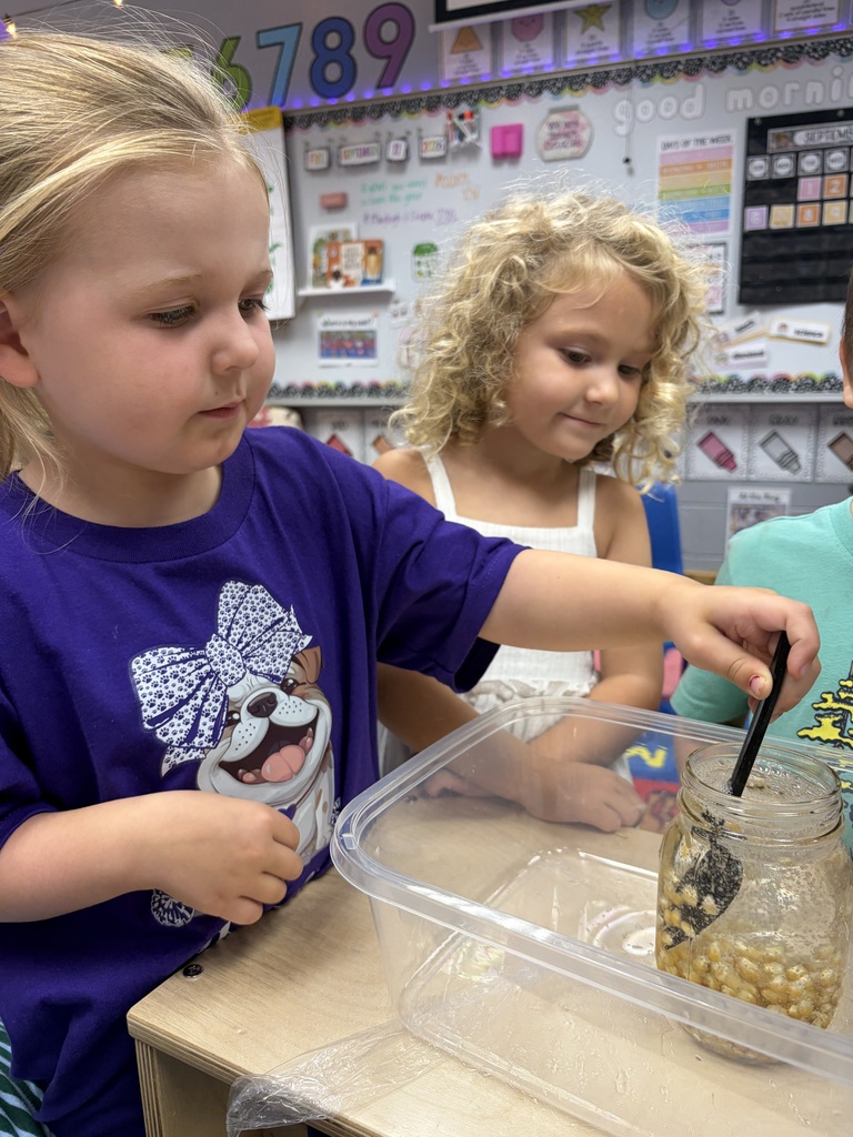Two young girls are doing an activity with a jar of beans, one in a blue shirt holding a stick.